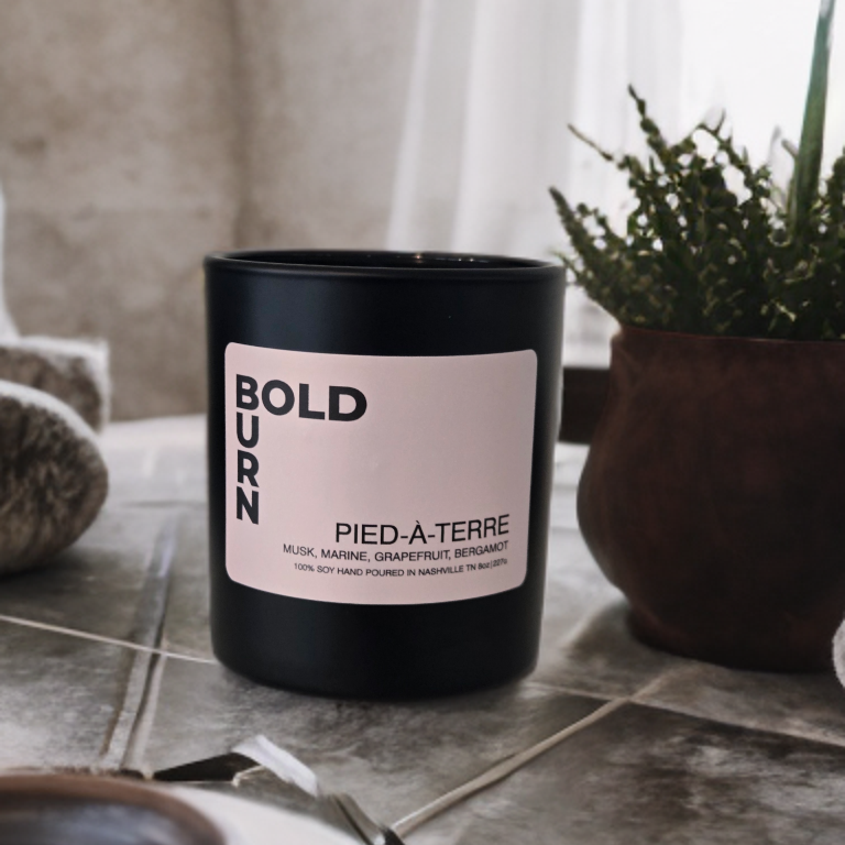 A matte black candle jar with a pale pink label reading 'BOLDURN' and 'PIED-à-TERRE' sits on a stone-tiled surface in a styled interior setting with a potted plant and soft natural light.