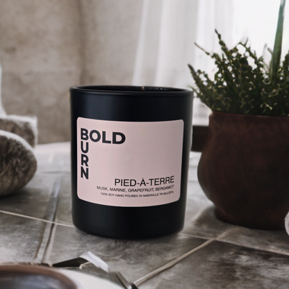 A matte black candle jar with a pale pink label reading 'BOLDURN' and 'PIED-à-TERRE' sits on a stone-tiled surface in a styled interior setting with a potted plant and soft natural light.