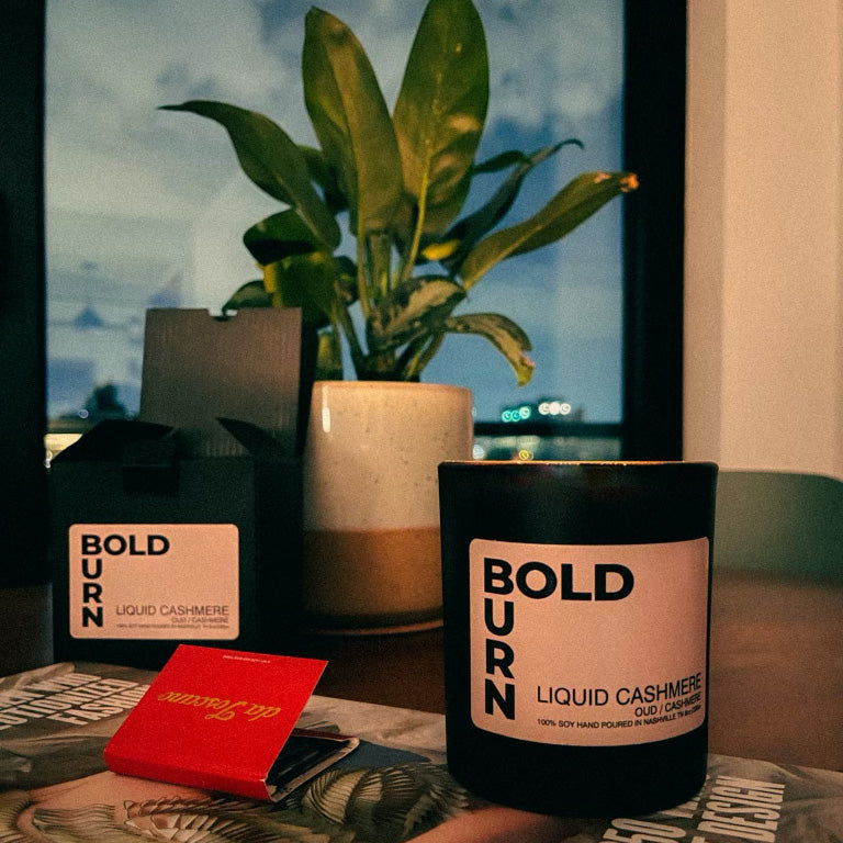 BoldBurn Liquid Cashmere two wick candle on a table with matches, candle gift box and plant
